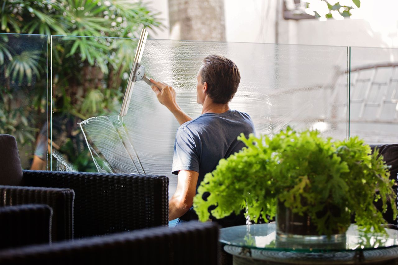 Un homme nettoie une barrière en verre à l’extérieur avec une raclette, entouré de verdure.